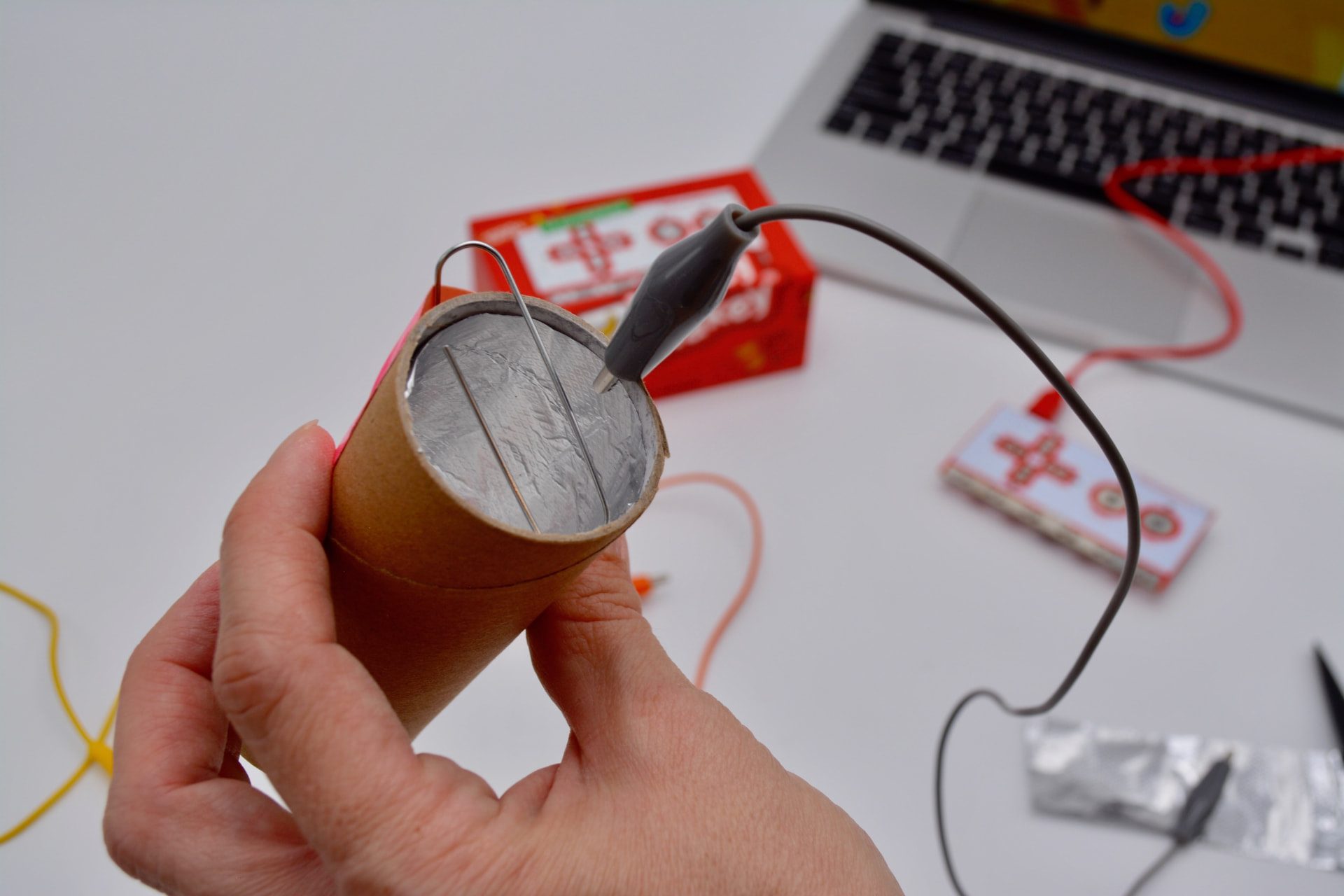 A hand holding a cardboard tube wrapped with foil and paperclips, wired with alligator clips to a Makey Makey for a physics conductivity experiment