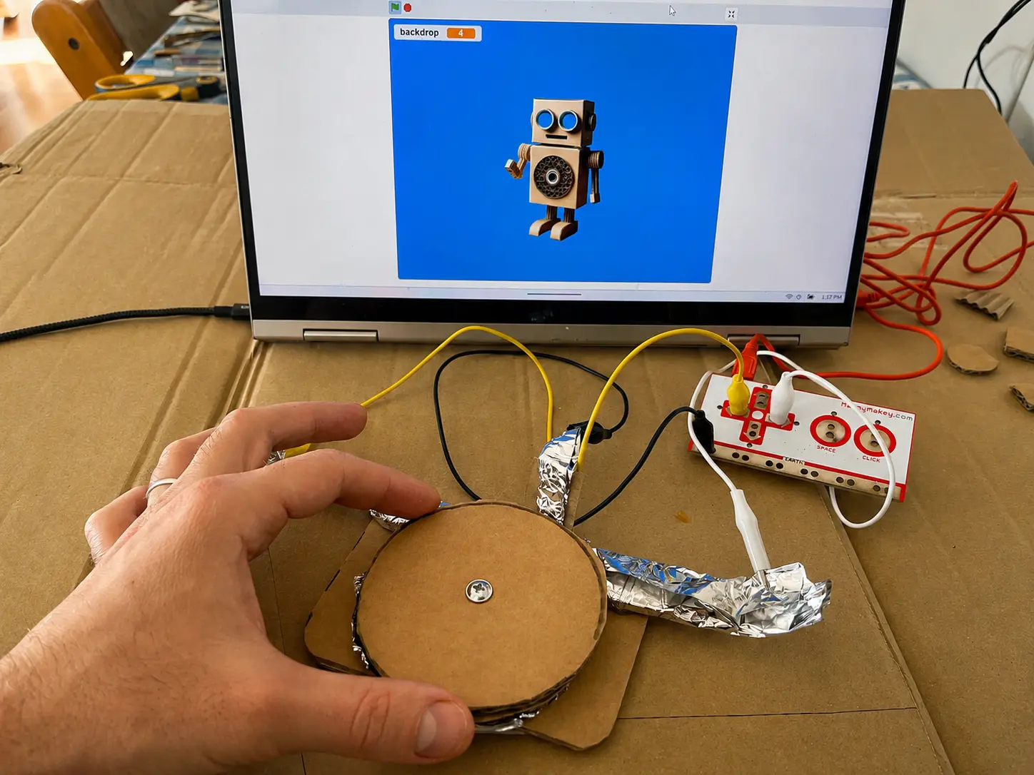 A hand pressing a round cardboard button wrapped in foil, wired to a Makey Makey controlling a robot in Scratch