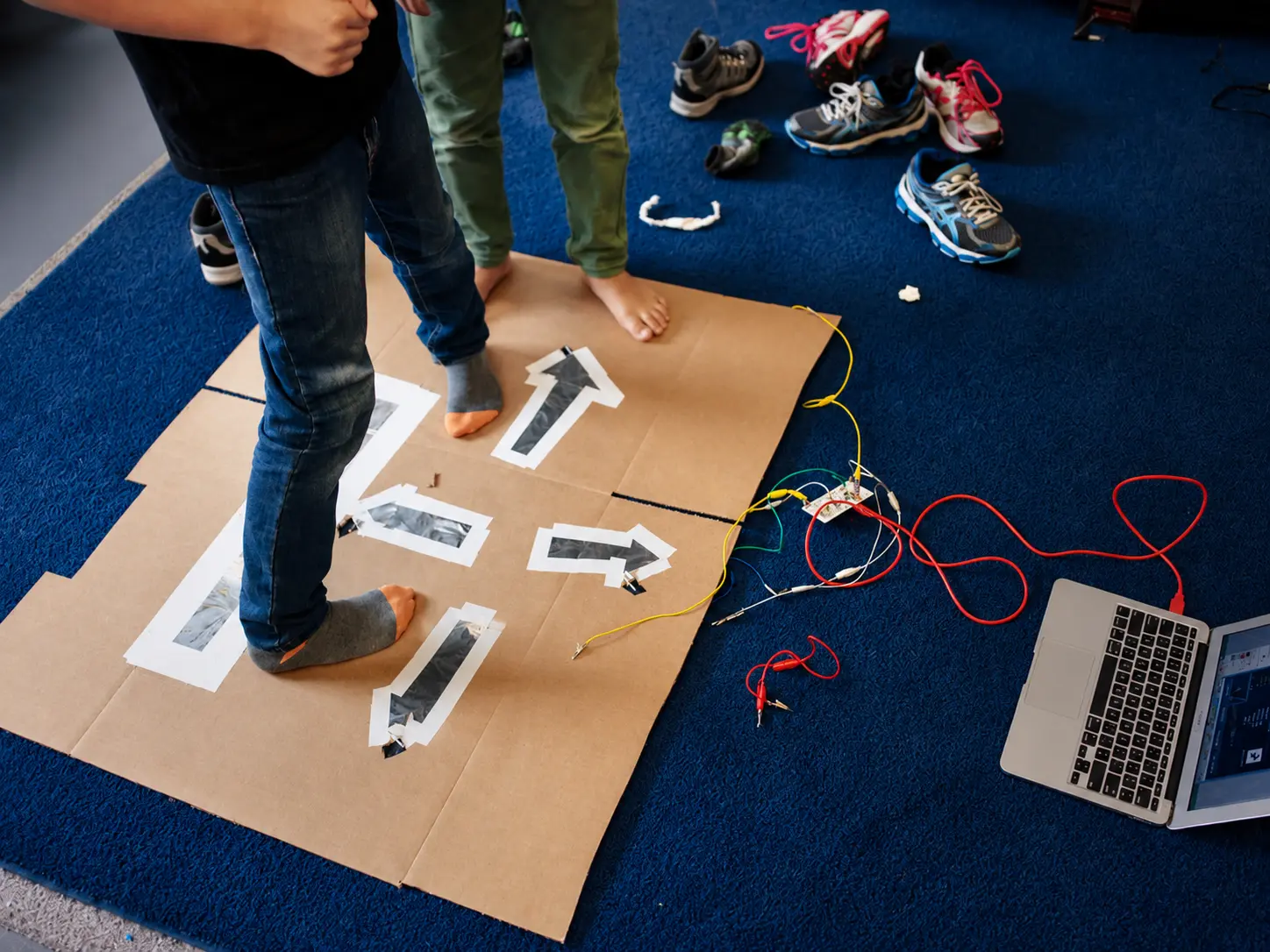 Two kids in socks stepping on a cardboard dance pad with foil arrow pads wired to a Makey Makey and a laptop