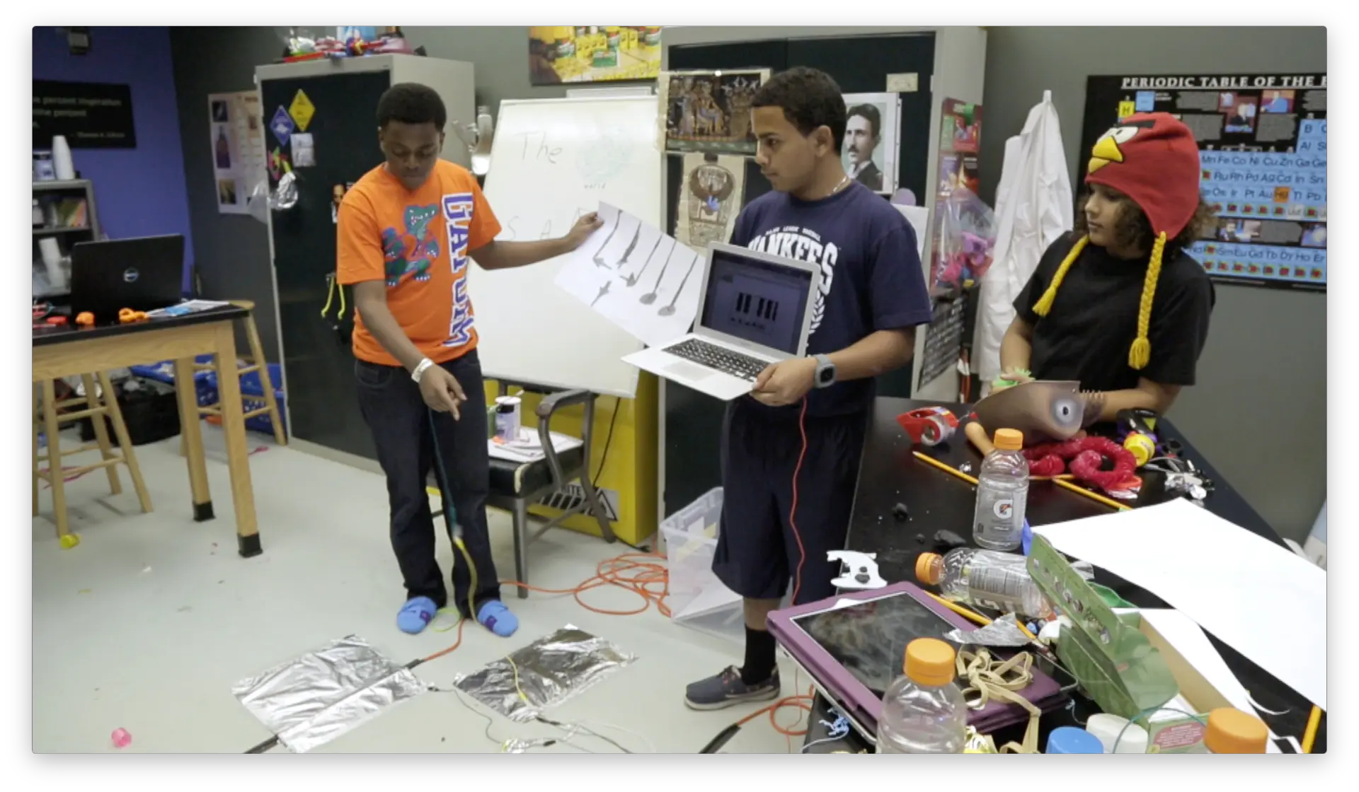 Three students collaborating with a Makey Makey invention, laptop, and conductive materials in a classroom