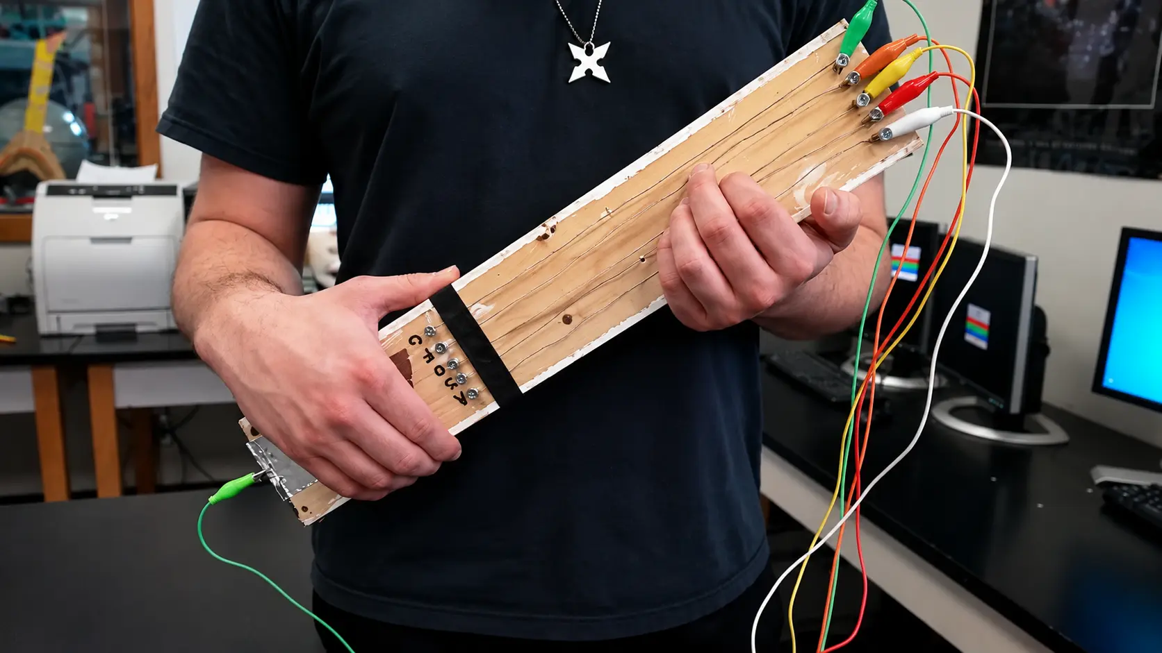 A person holding a plank-of-wood guitar with metal screws as frets, wired with alligator clips to a Makey Makey