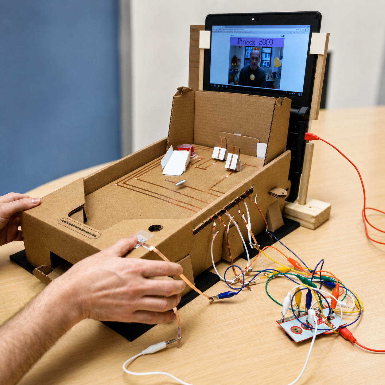 Hands playing a cardboard pinball box with copper tape targets wired by alligator clips to a Makey Makey and a laptop