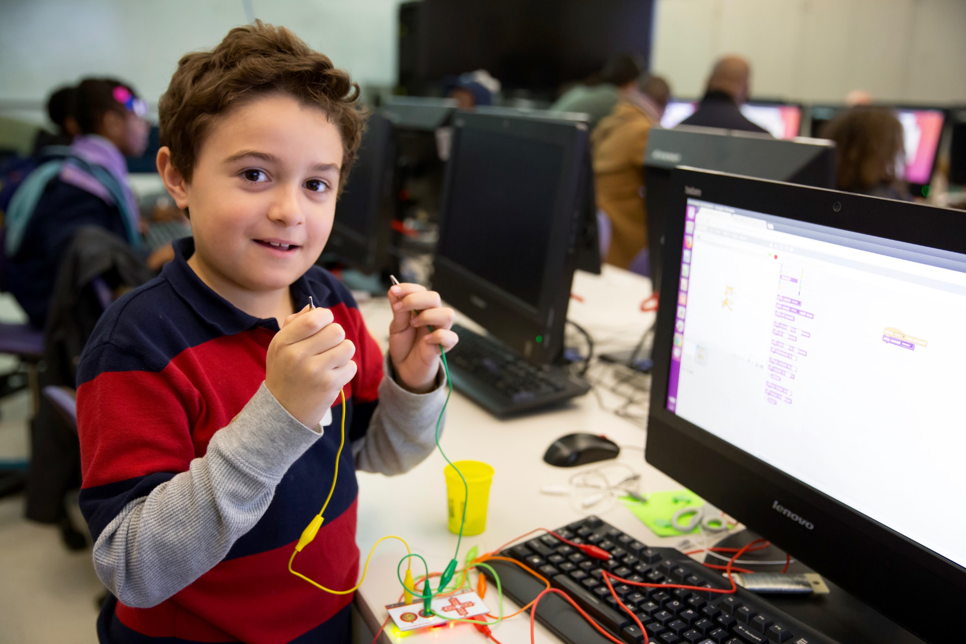 A young student smiling while holding alligator clips connected to a Makey Makey board in a computer lab
