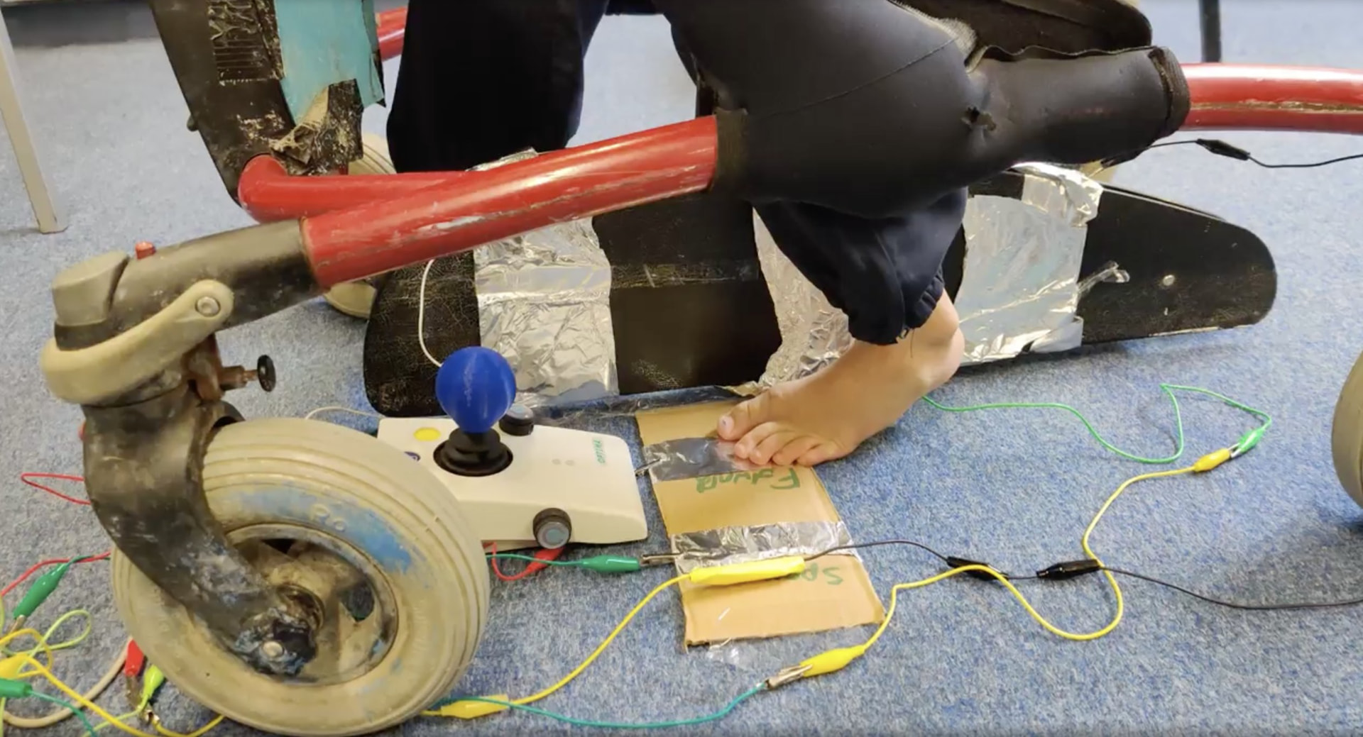 Student using a custom foot-operated Makey Makey controller with a wheelchair nearby