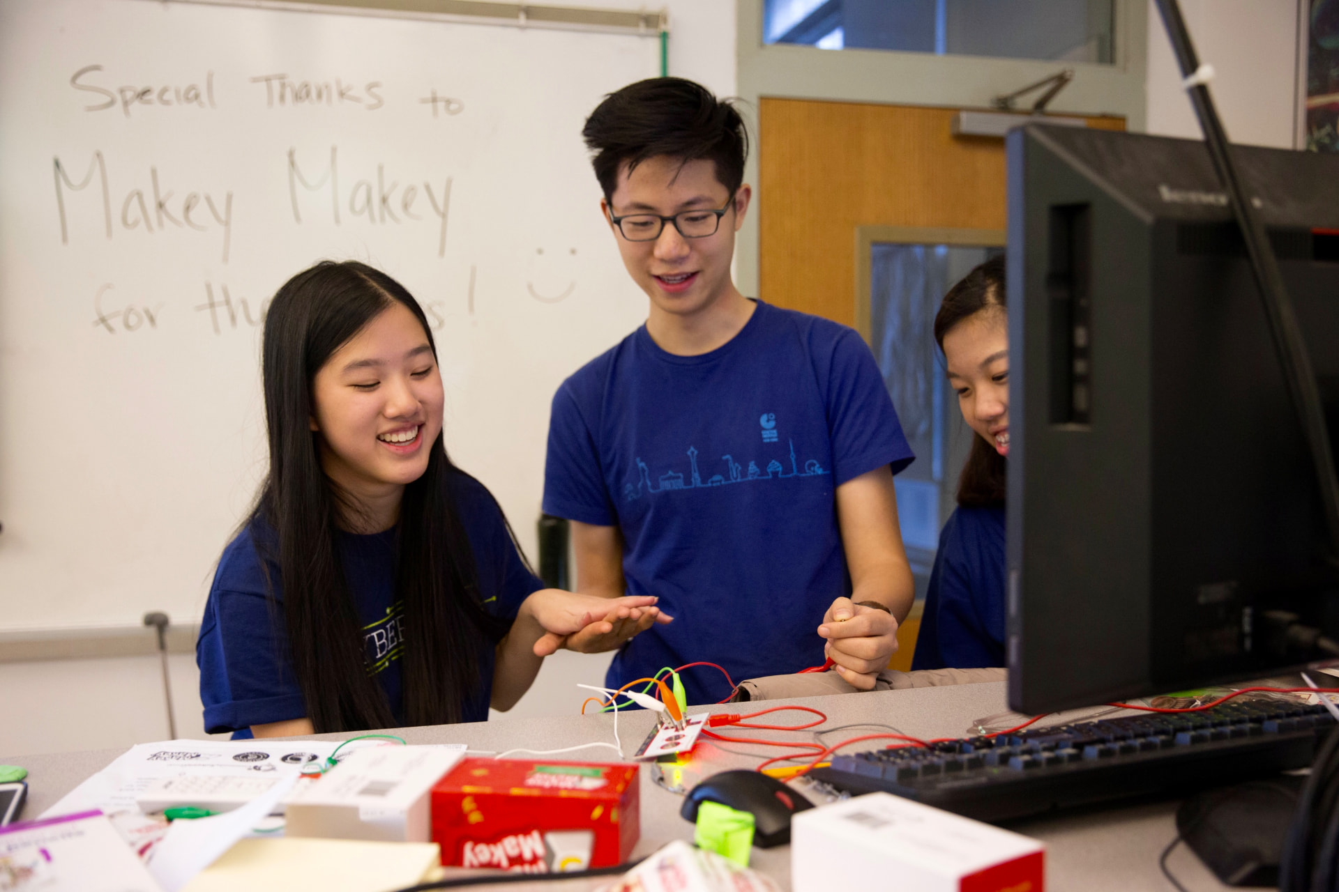 Three students smiling while collaborating on a Makey Makey project in a classroom