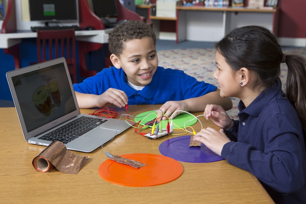 Two students collaborating with a Makey Makey invention connected to a laptop