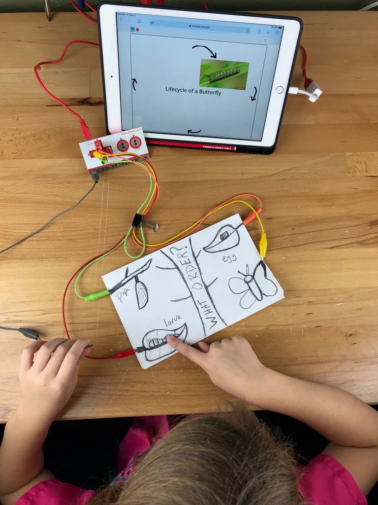 First grader touching a hand-drawn butterfly lifecycle diagram wired to a Makey Makey