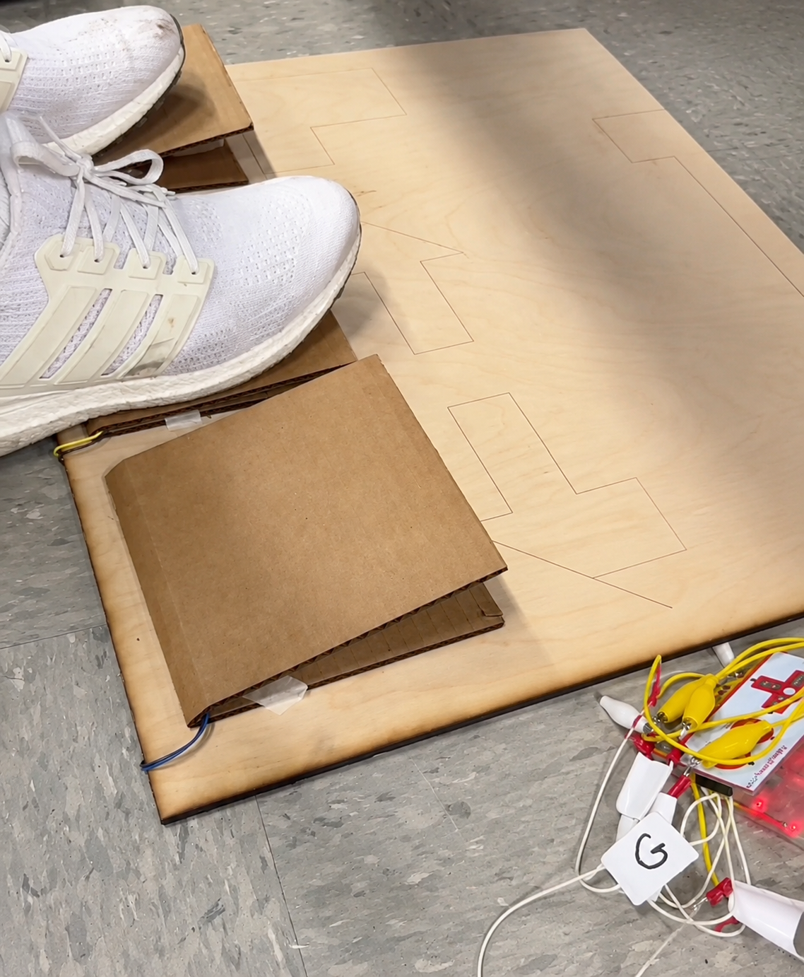 Sneakers pressing cardboard foot pedals mounted on a plywood board wired with alligator clips to a Makey Makey