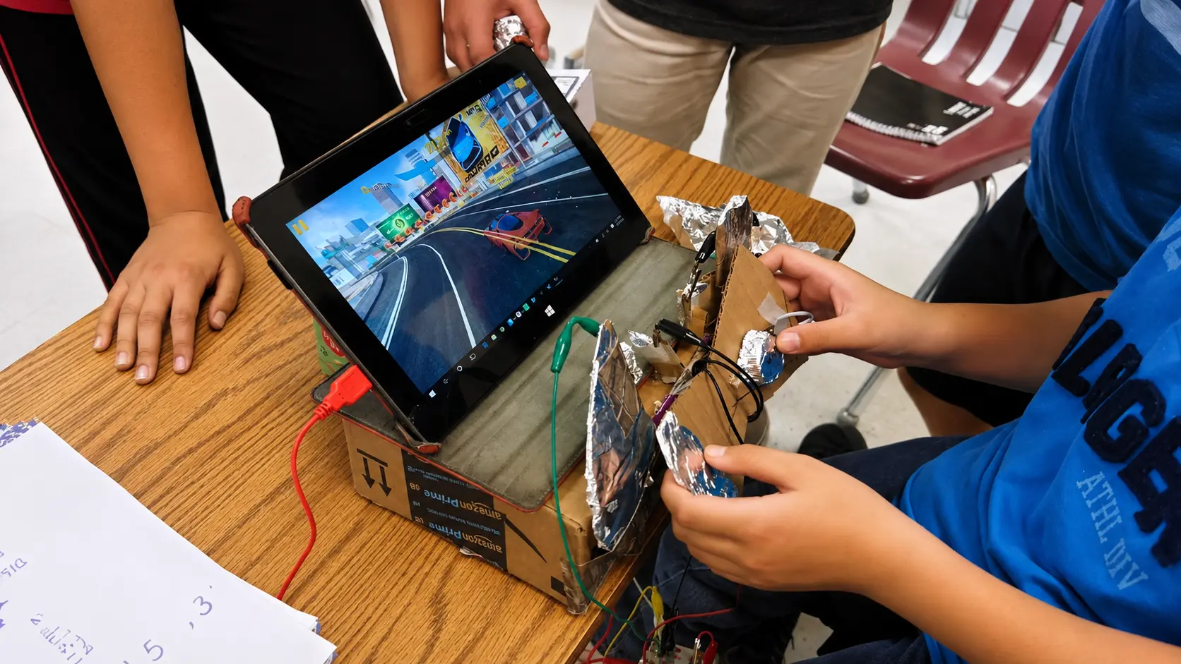 Students playing a racing game with a cardboard-and-foil steering controller wired to a Makey Makey and a tablet