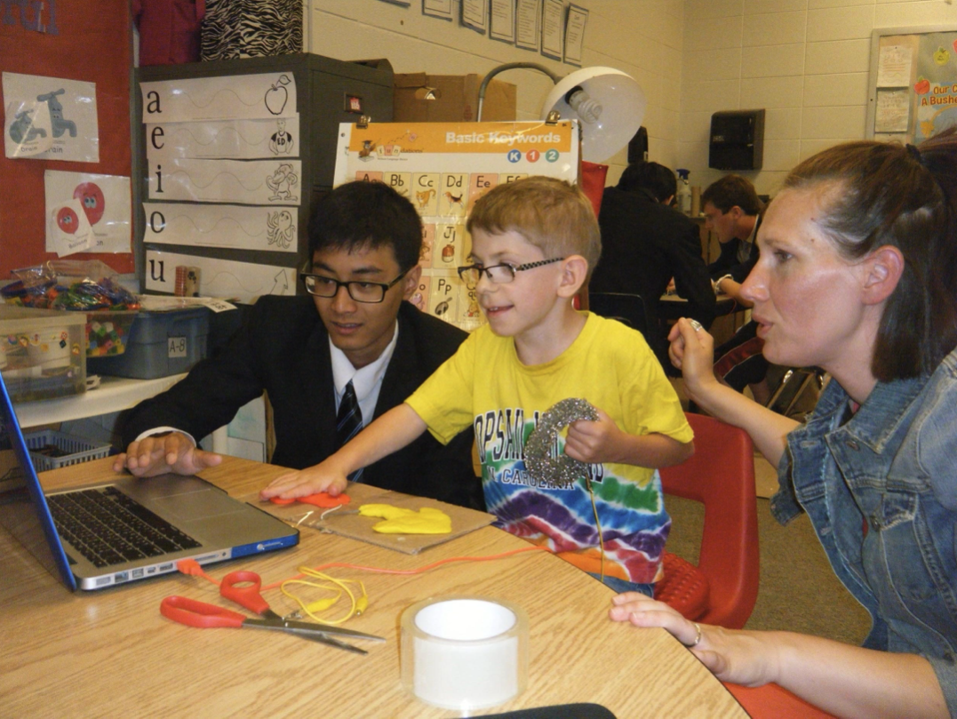 Two students and an adult collaborating with a Makey Makey kit, laptop, and conductive materials in a classroom