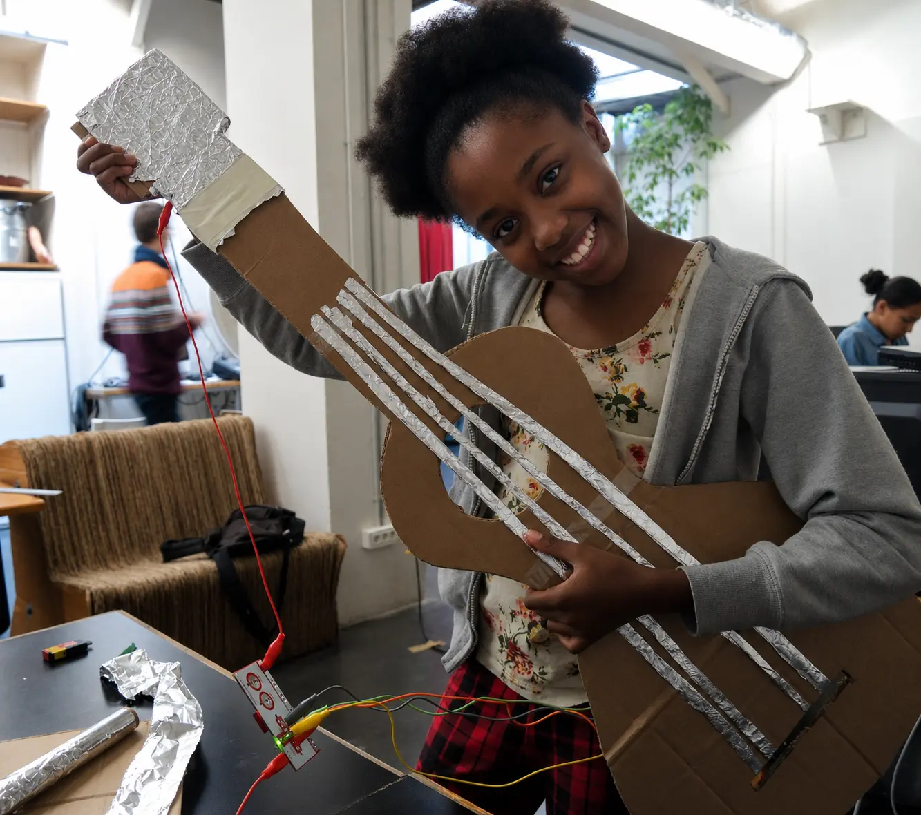 A student smiling while holding a cardboard guitar with aluminum foil strings connected to a Makey Makey