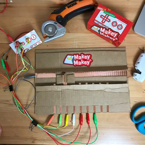 A cardboard slider switch with copper tape contacts and colorful alligator clips wired to a Makey Makey on a workbench